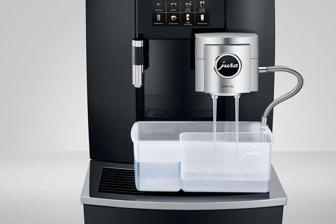 Jura Giga X8c bean-to-cup coffee machine performing automated cleaning cycle with dual spouts and milk system cleaner container.